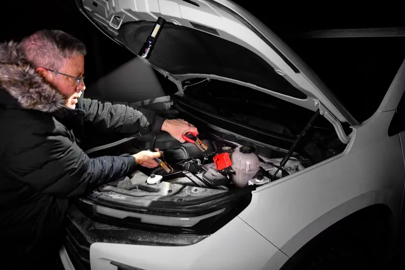 A man using the Lumen Force flashlight magnetically attached to a raised car hood, lighting the engine bay hands-free while connecting jumper cables at night.